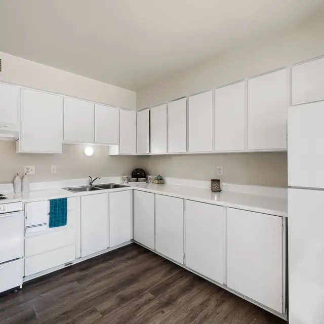 A modern kitchen featuring white cabinets, a white refrigerator, and a white stove. The countertop is light and there is a teal towel hanging from the oven. The floor is made of wood.
