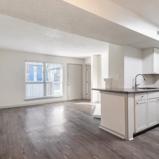 Interior of a spacious and modern apartment with light-colored walls, a kitchen area featuring white cabinets, and a large living area with a window.