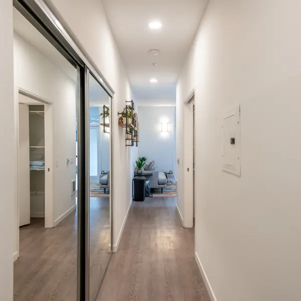 A long hallway with smooth walls and a wooden floor, featuring mirrored closet doors on the left and a doorway leading to a living area at the end.