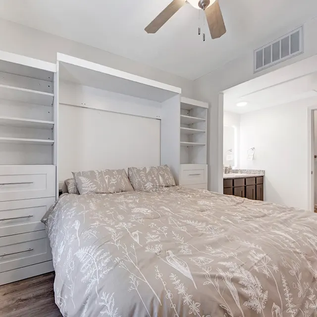 Modern Bedroom Design A modern bedroom featuring a king-size bed with light-colored bedding, built-in white storage shelves on either side, and a ceiling fan above. The room has wooden flooring and a doorway leading to a bathroom.