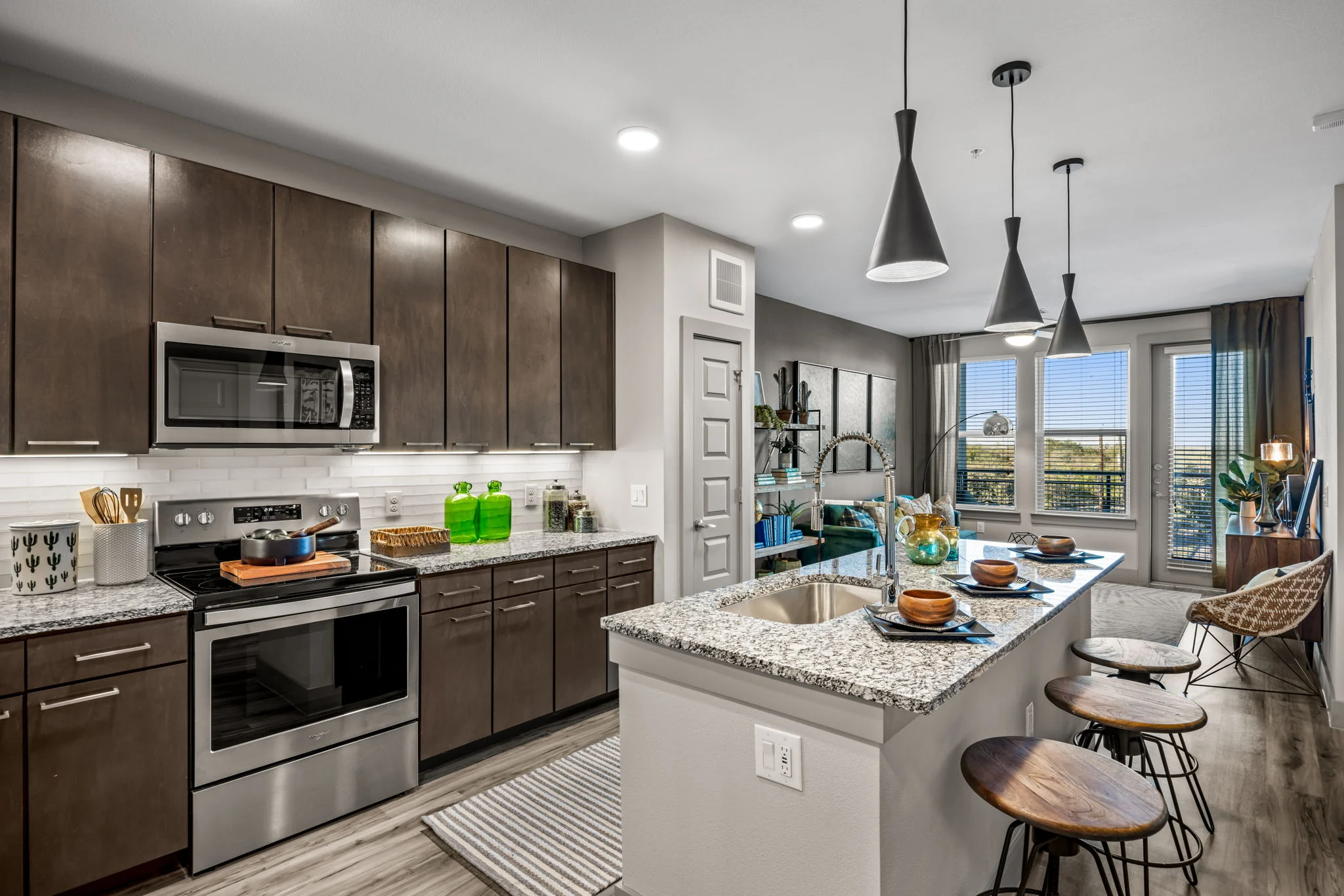 A modern kitchen featuring dark wood cabinets, stainless steel appliances, and granite countertops. An island with bar stools and decorative items is in the center, with a large window letting in natural light.