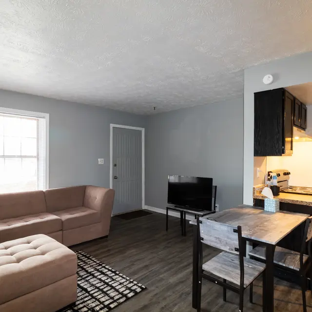 A cozy living room with a modern design featuring a light gray color scheme, a beige sectional sofa, a matching ottoman, and a wooden dining table with chairs. A television is positioned near the table, and large windows let in natural light.