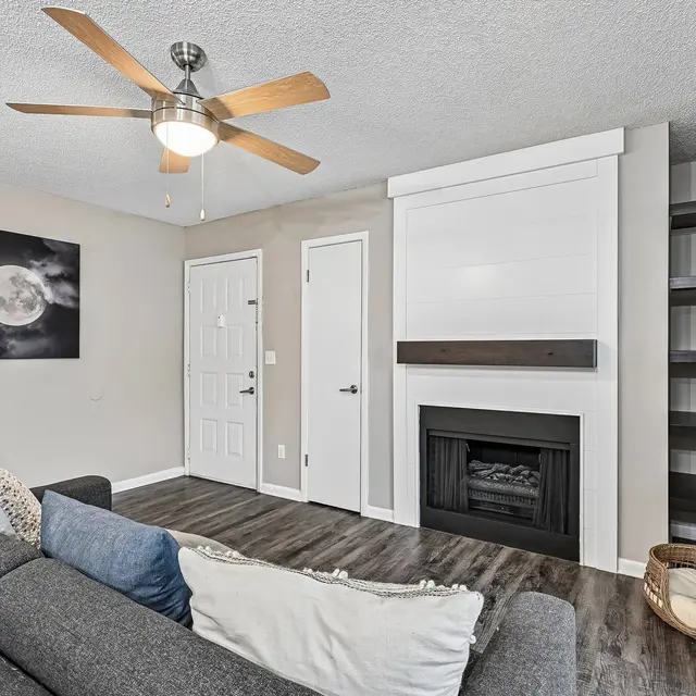 Cozy Living Room with Fireplace A cozy living room featuring a gray sofa with decorative pillows, a ceiling fan, and a wall-mounted fireplace. A painting of a moon is hung on the wall, and there are doors leading to other rooms.
