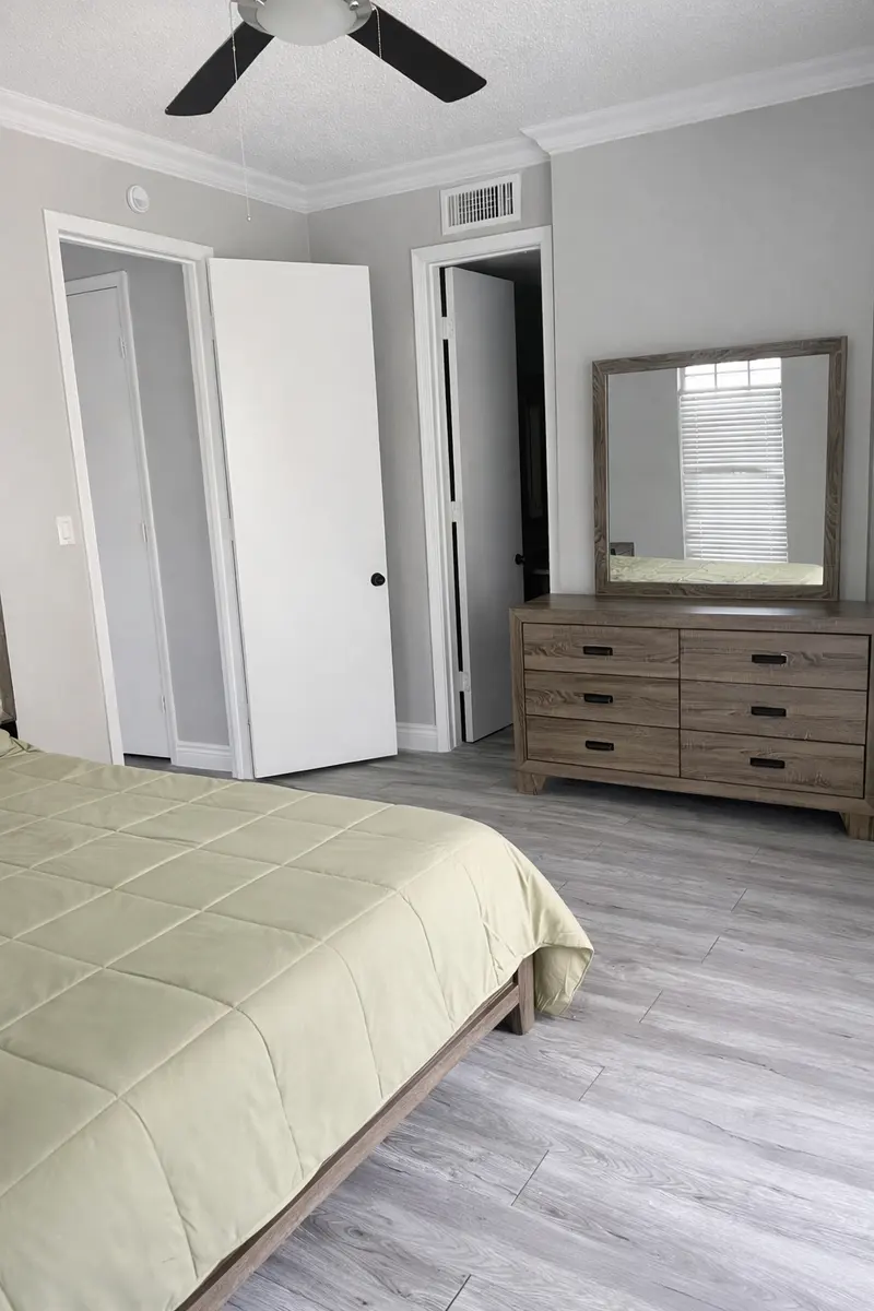 A modern bedroom featuring a bed with a light green quilt, a wooden dresser, a mirror, and two doors leading to adjacent rooms. The floor is light-colored and the walls are painted in a soft gray. A ceiling fan is visible.