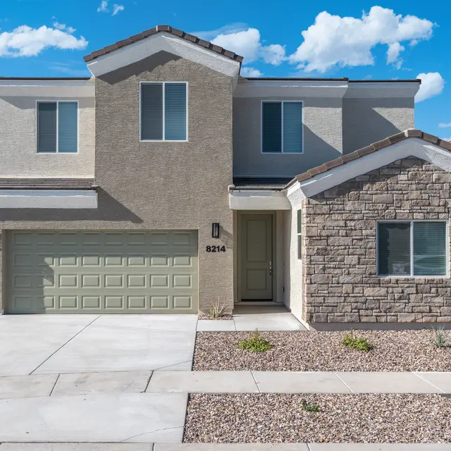 A modern two-story house featuring a combination of stucco and stone exterior, with a green garage door and multiple windows under a blue sky with fluffy clouds.