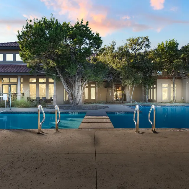 A tranquil view of a swimming pool surrounded by trees and a residential building in the background during sunset.