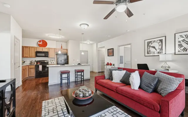 A stylish modern apartment living room featuring a red sofa with decorative pillows, a coffee table with ornamental bowls, and a kitchen area with bar stools and contemporary appliances.