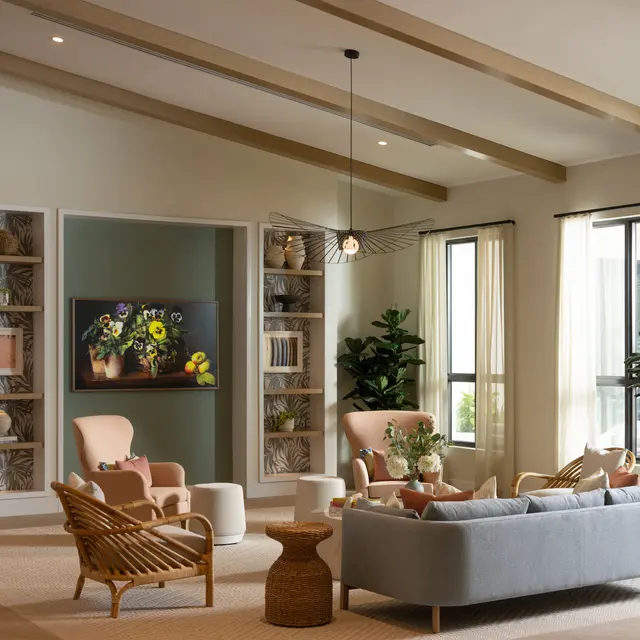 A modern living room featuring natural light, a large sofa, stylish chairs, and decorative plants. The room has built-in shelves with art and books, and a TV displaying floral artwork.