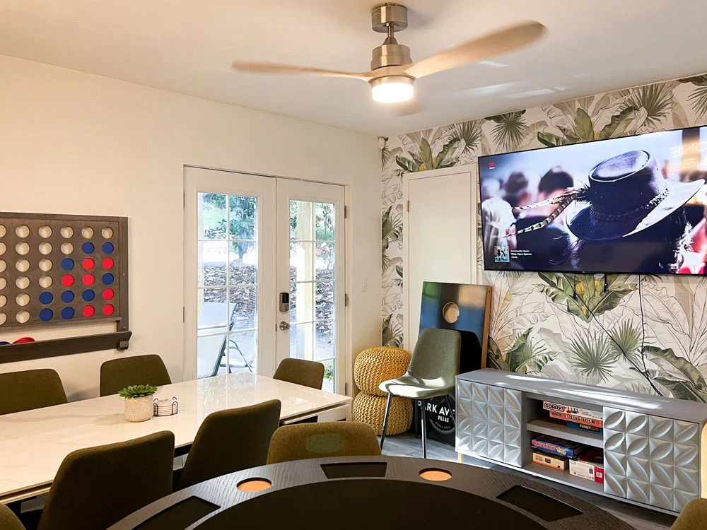 A modern meeting room with a round black table, green chairs, and a large TV mounted on the wall. The wall features tropical leaf wallpaper and a large game board on the left. Large glass doors lead to an outdoor area.
