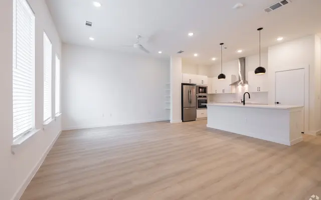 A spacious, modern apartment interior featuring an open floor plan with a kitchen and living area. Large windows let in natural light. The kitchen has stainless steel appliances and pendant lights above the counter.