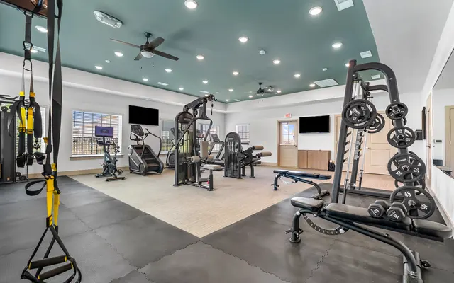 A spacious gym interior featuring various exercise equipment like weight machines, free weights, and a cardio area, with large windows and a ceiling fan.