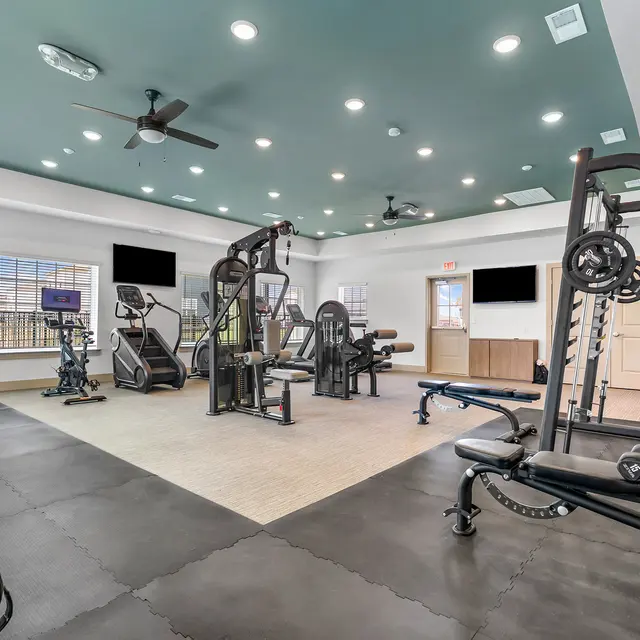 A spacious gym interior featuring various exercise equipment like weight machines, free weights, and a cardio area, with large windows and a ceiling fan.
