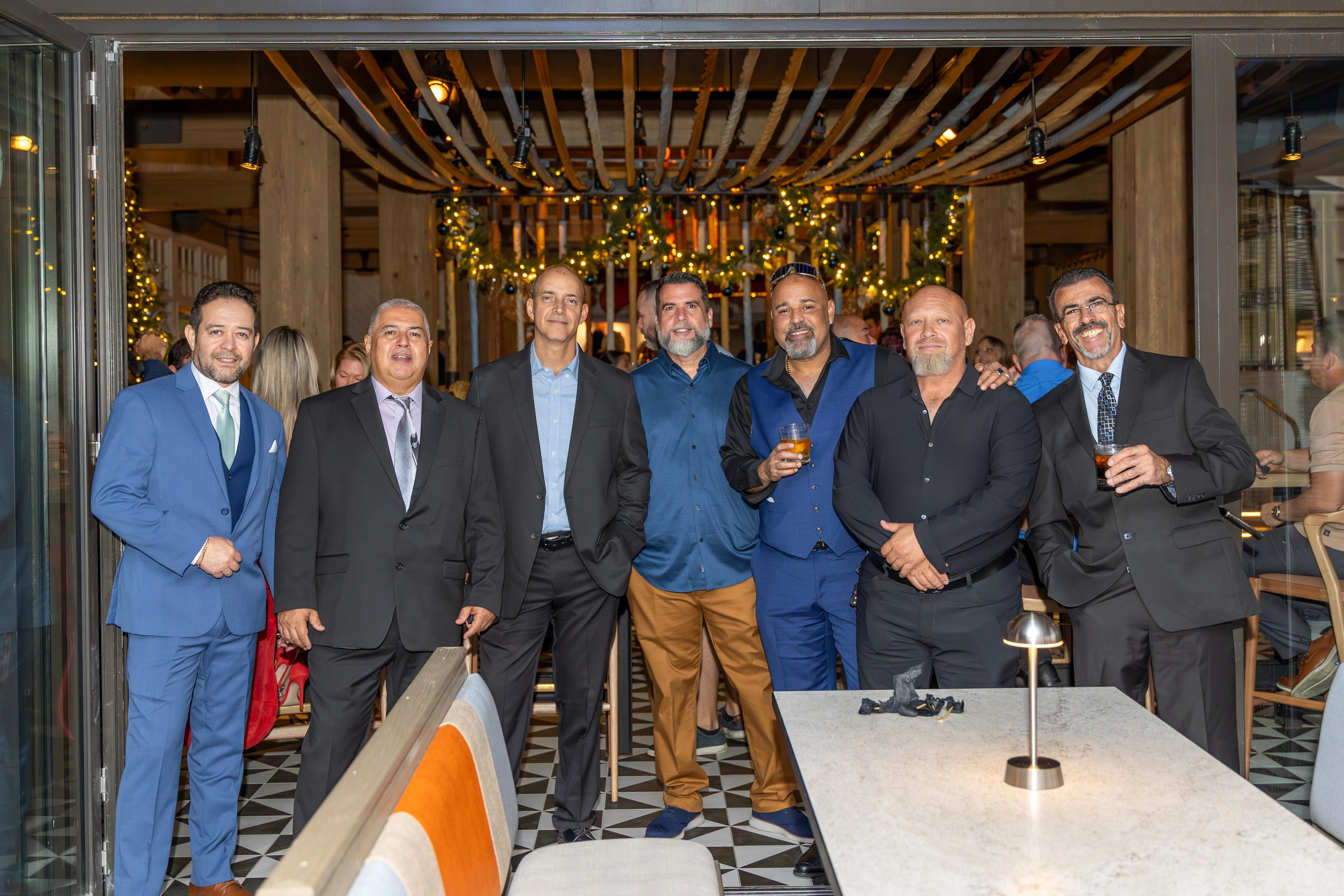 A group of seven men standing together in a stylish restaurant, smiling and posing for the photo. The decor features modern furniture and festive lighting.
