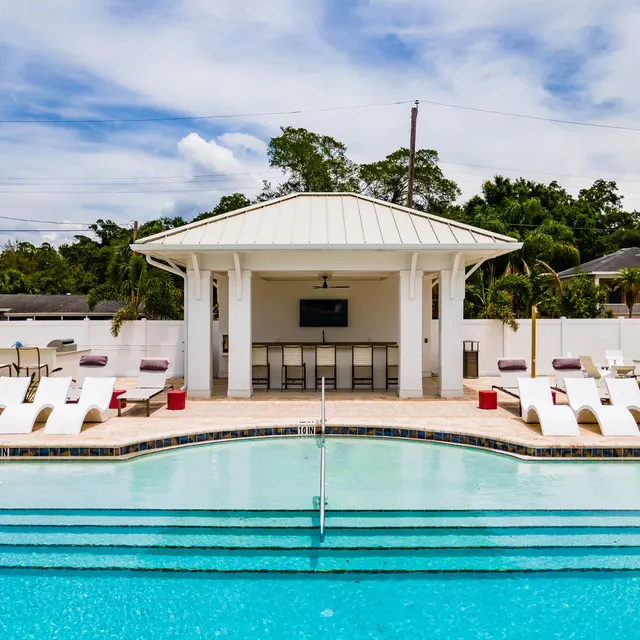 A spacious pool area featuring a light-colored cabana, lounge chairs, and a clear blue pool under a bright sky.