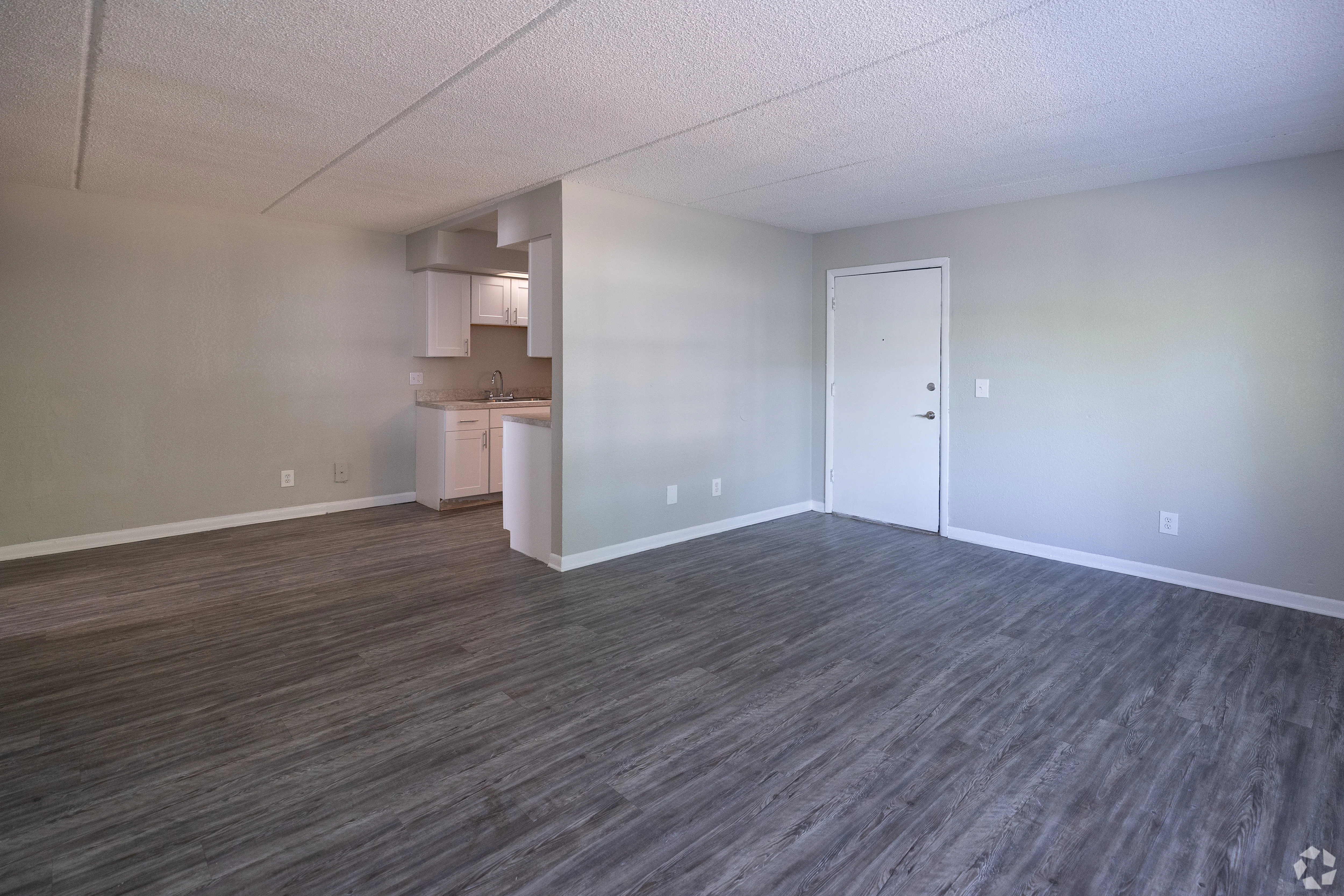 Modern Apartment Living Space A spacious, empty living area with light-colored walls and gray laminate flooring. There is a kitchen area visible in the background with white cabinets. A white door is seen on the right side of the image.