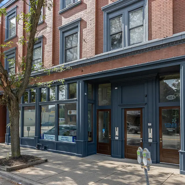 Exterior view of a historic multi-story building with brick facade, large windows, and a sidewalk lined with trees.