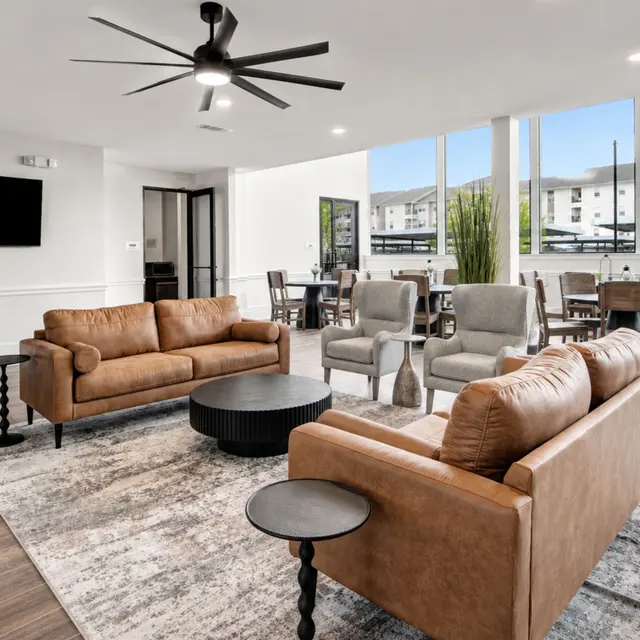 Spacious Community Lounge Area A modern community lounge area with comfortable seating, a ceiling fan, and large windows letting in natural light. The space includes brown leather couches, armchairs, and tables arranged for social gatherings.