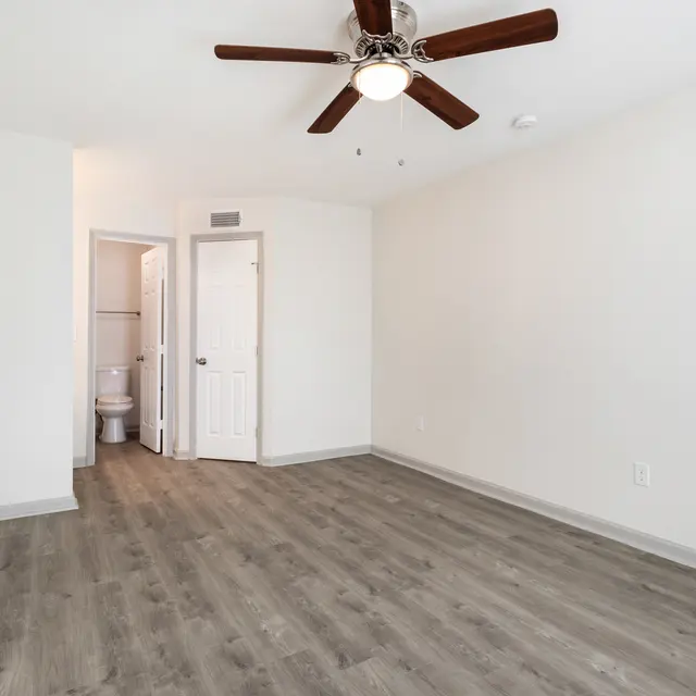 An empty room featuring a ceiling fan, light-colored walls, and laminate flooring, with a bathroom visible in the background through an open door.