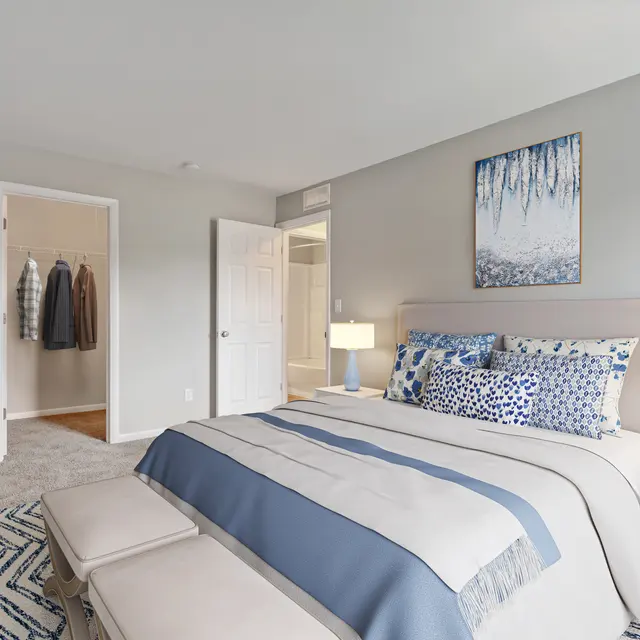 A modern bedroom featuring a large bed with blue and white bedding, two bedside lamps, and a piece of artwork on the wall. There is a small tree and a carpet on the floor, along with an open closet visible in the background.