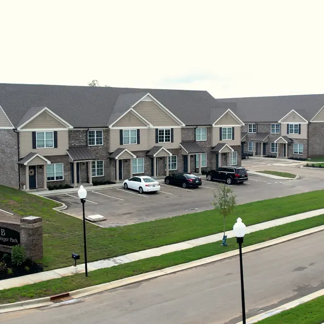 Aerial view of a residential apartment complex featuring brick buildings with multiple units. The foreground shows a landscaped area with flowers and light posts, while the parking lot displays several cars. The sky is overcast.