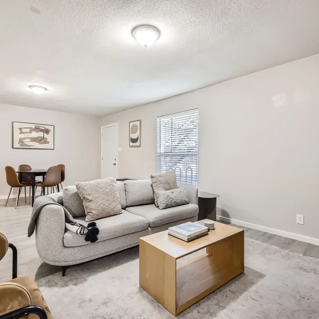 Cozy living room featuring a gray sofa, wooden coffee table, and dining area in the background.