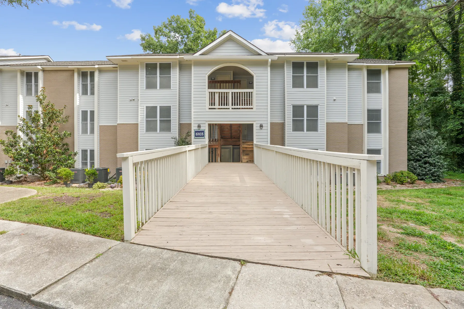 Exterior view of a multi-story apartment building with a wooden ramp leading to the entrance, surrounded by green landscaping and trees.