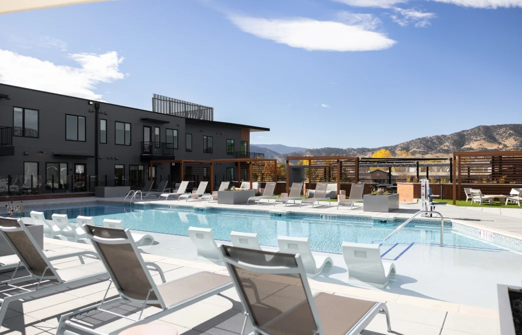 Luxury Apartment Pool Area An outdoor swimming pool surrounded by lounge chairs, with a modern apartment building in the background and mountains in the distance under a clear blue sky.