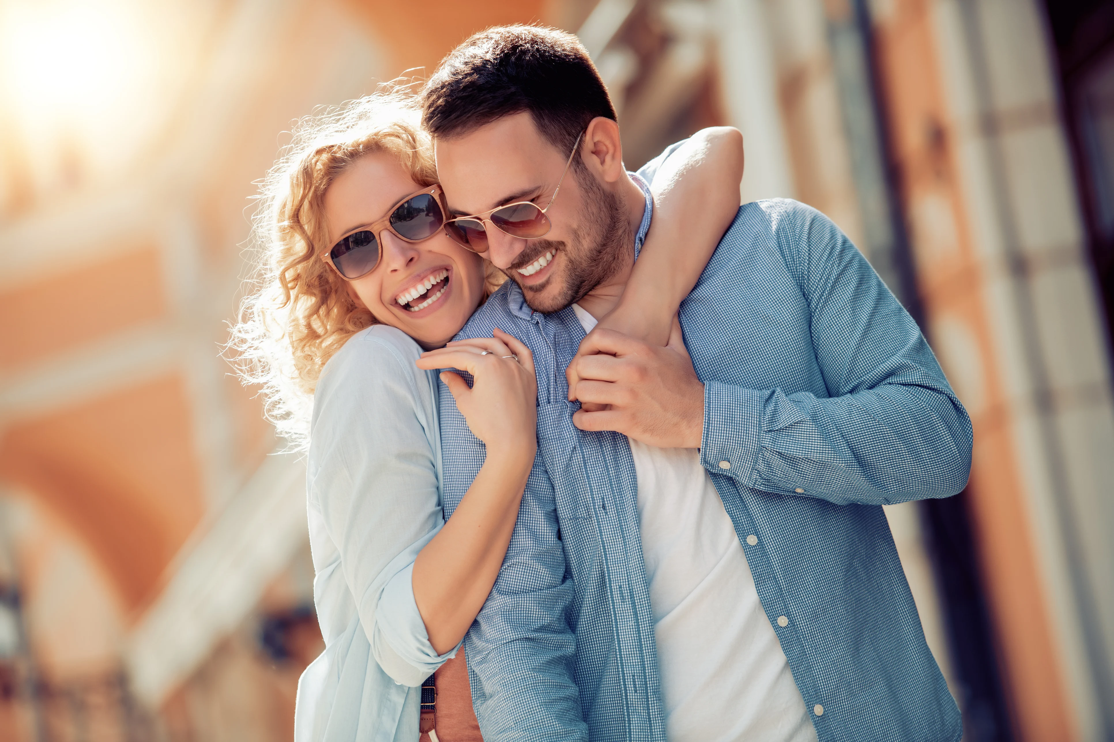 Happy Couple Embracing in Sunshine A joyful couple embracing outdoors, both wearing sunglasses and smiling. The sun is shining in the background.