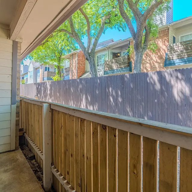 A view from a small balcony overlooking a wooden fence and nearby buildings, with greenery visible in the distance.