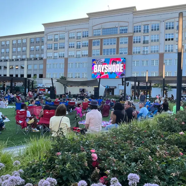 A large crowd of people sitting in colorful lawn chairs on a grassy area, with a large screen displaying 'BAYSHORE' in the background, and a modern building behind.