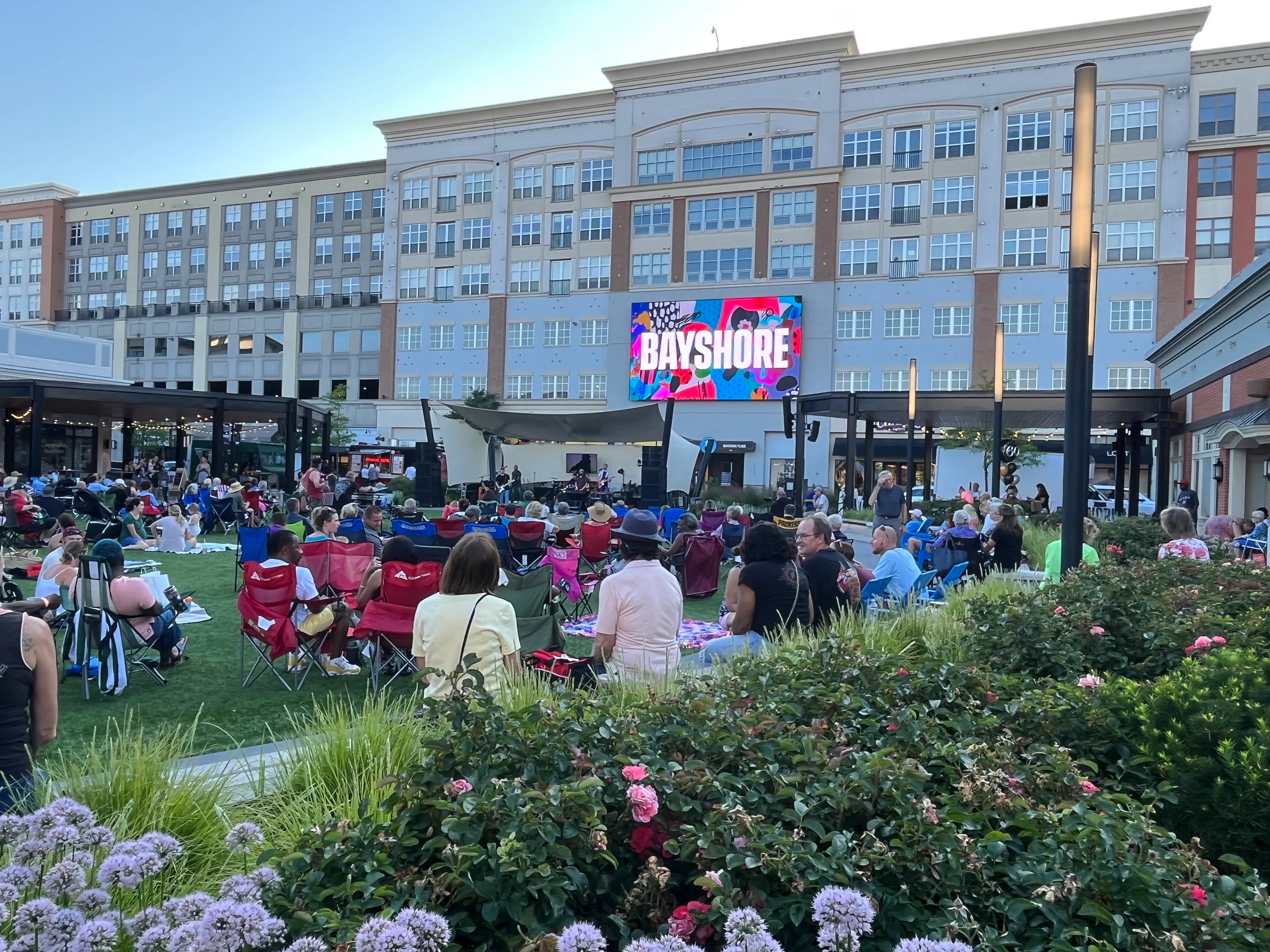 A large crowd of people sitting in colorful lawn chairs on a grassy area, with a large screen displaying 'BAYSHORE' in the background, and a modern building behind.