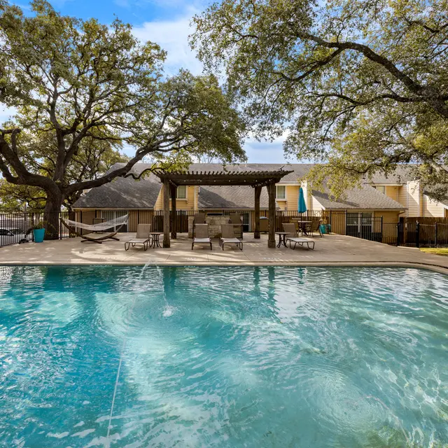 A serene swimming pool area with lounge chairs and a pergola surrounded by large trees.