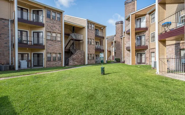 A view of an apartment complex featuring three buildings surrounding a green lawn area. Each building has balconies and brick exterior. The lawn is neatly maintained with a few plants and walkways visible.