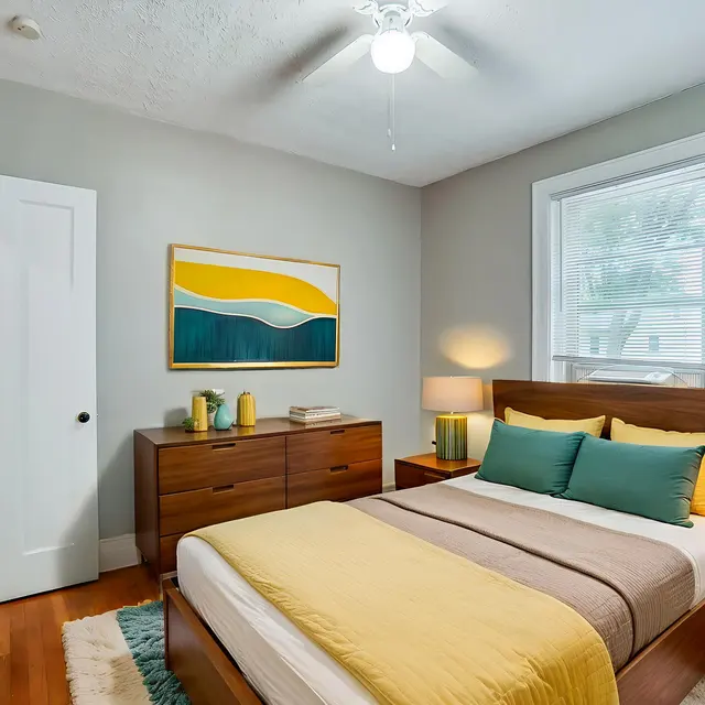 A cozy bedroom featuring a bed with yellow and teal accents, wooden furniture, and large windows allowing natural light in.