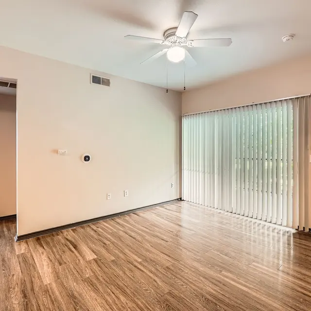 A spacious living room with wooden flooring, a ceiling fan, and large sliding glass doors leading to a patio, with an open doorway visible to another room.