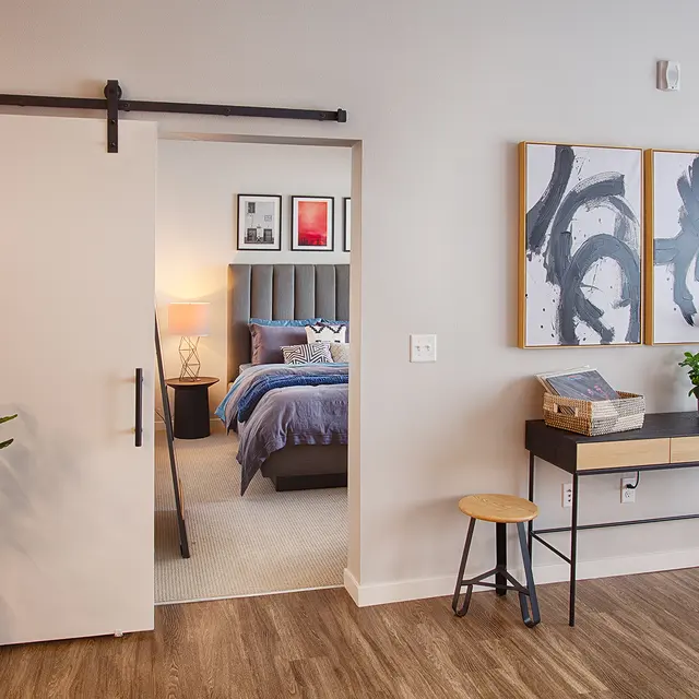 Modern interior with a sliding door, a small desk, a stool, and a glimpse of a bedroom featuring blue bedding through the door.