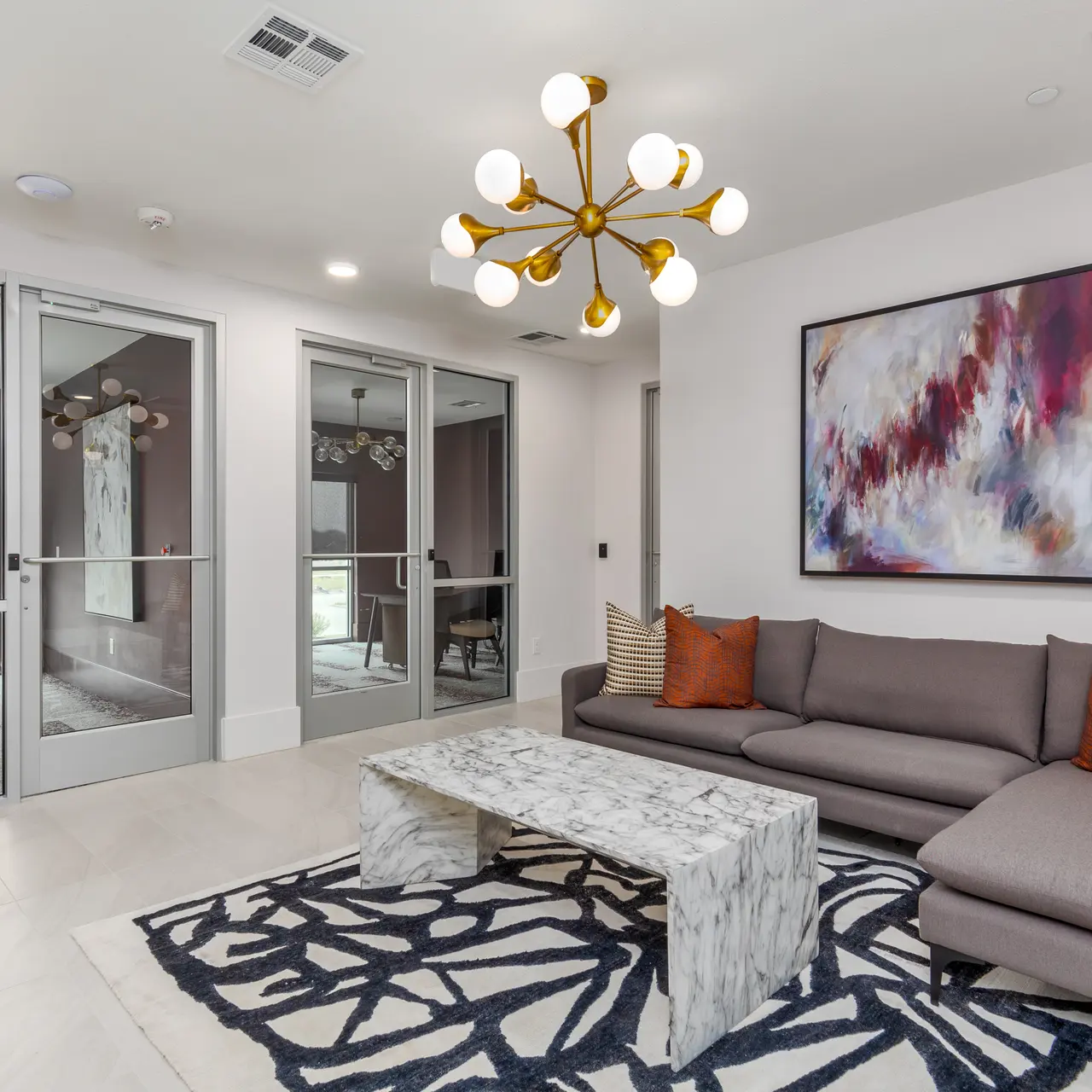 Stylish Modern Living Room A modern living room with a gray sectional sofa, an elegant marble coffee table, and a vibrant abstract painting on the wall. The floor is decorated with a geometric rug, and large glass doors lead to another room, allowing natural light to fill the space.