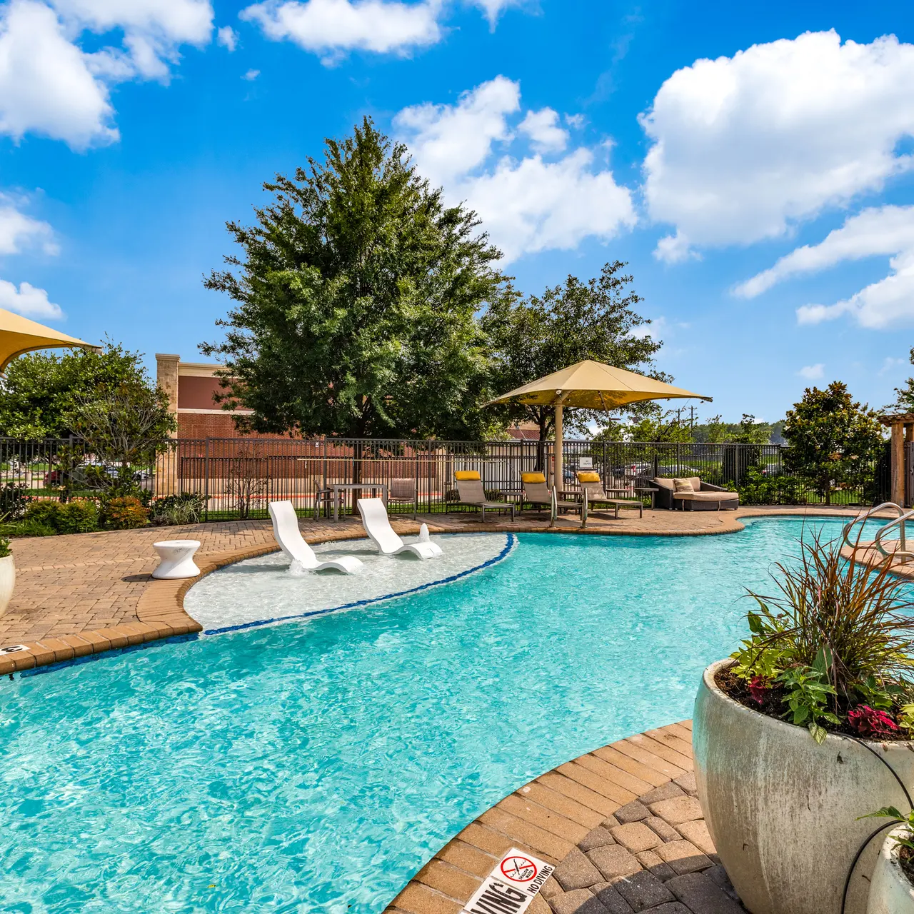 Sunny outdoor pool area with lounge chairs and umbrellas surrounded by greenery.