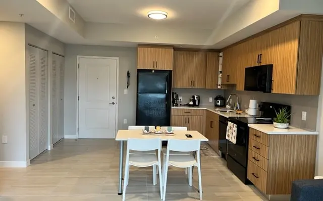 A modern kitchen and dining area featuring light wood cabinetry, a black refrigerator and stove, a small white dining table with two chairs, and creamy tile flooring.