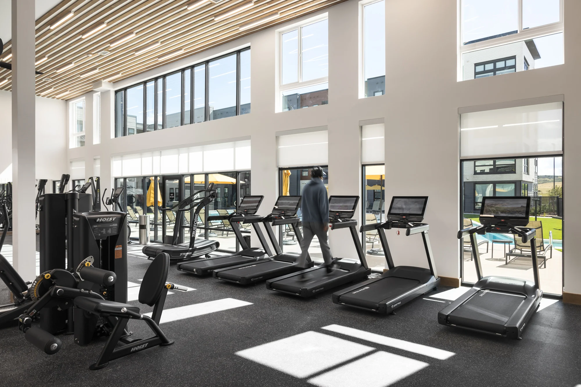 A modern gym interior with large windows letting in natural light. Several treadmills and exercise machines are visible, as well as a person exercising on one of the treadmills.