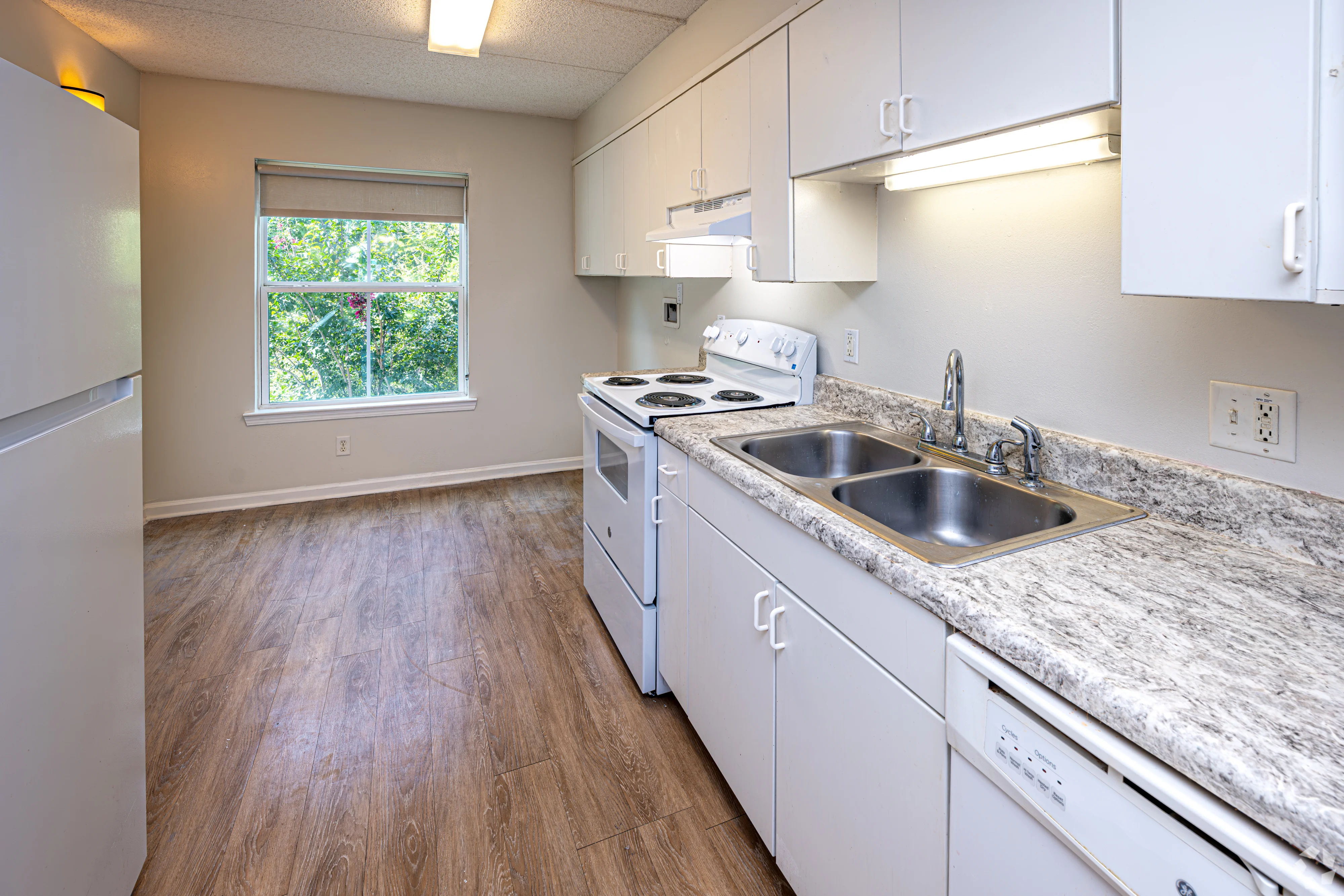 A modern kitchen featuring white cabinets, a gray countertop, and a view of greenery through a window. The appliances include a refrigerator, stove, and a double sink.