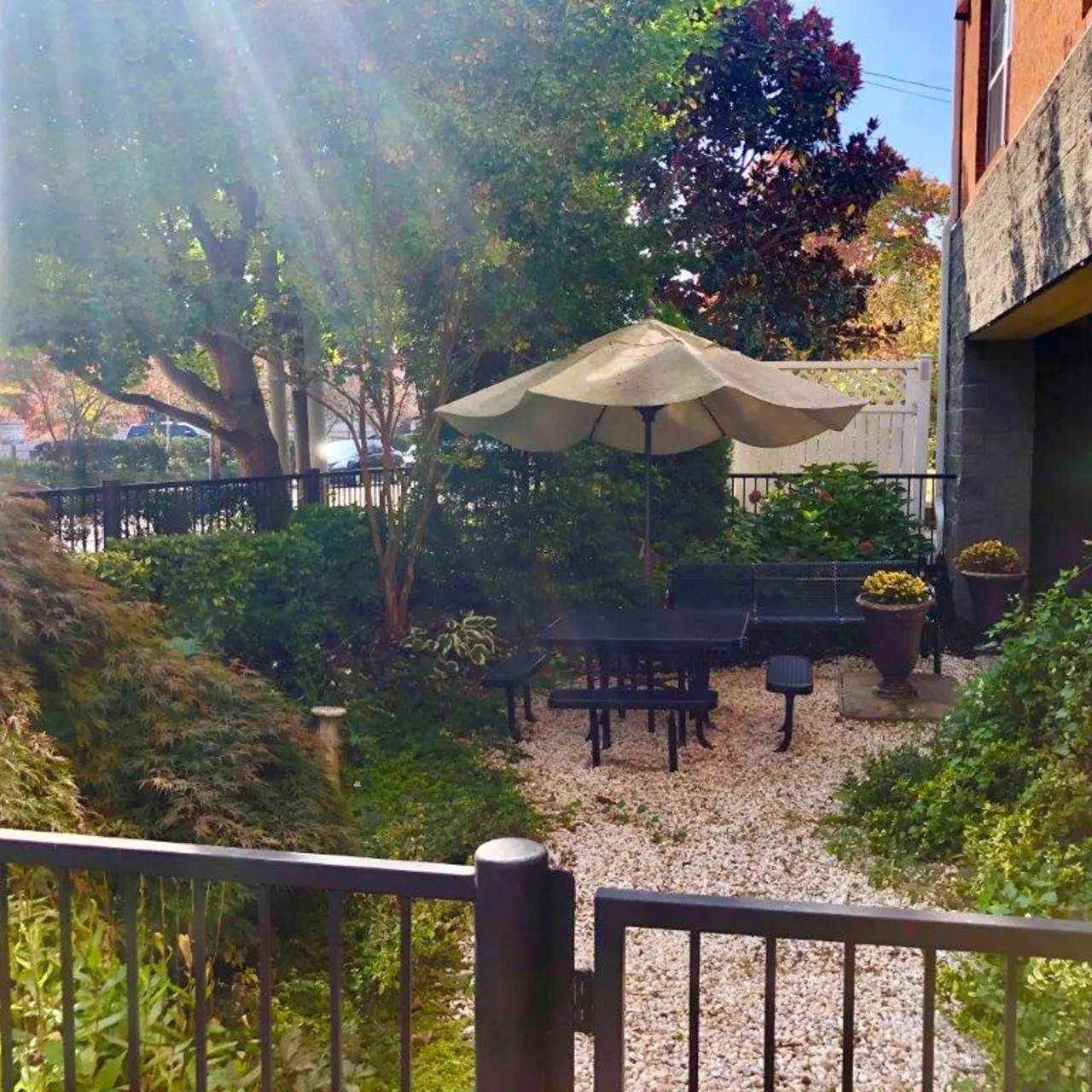 A tranquil garden area featuring a patio umbrella over picnic tables surrounded by greenery and decorative plants, enclosed by a low fence.