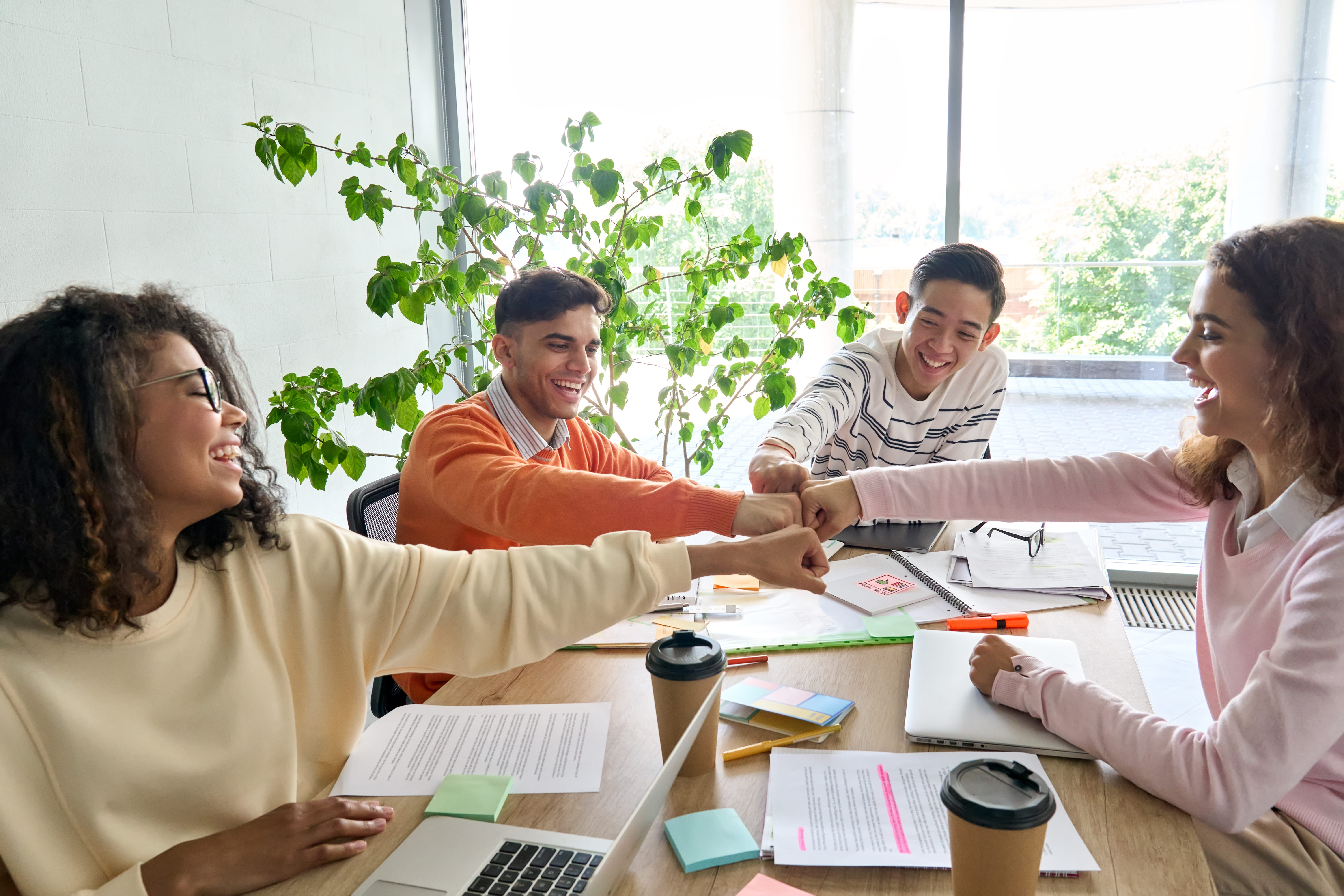 Team Collaboration Four young adults engaging in a fist bump while sitting around a table filled with notebooks and coffee cups, surrounded by indoor plants and large windows.