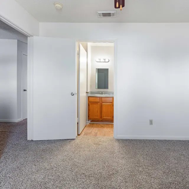 A view of an empty room with carpet, featuring an open door leading to a bathroom area with a vanity.