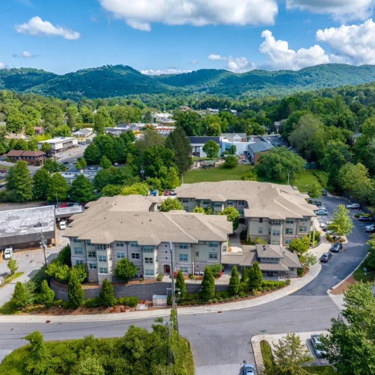 Aerial view of a residential area surrounded by green hills and trees, featuring modern buildings and a parking area.