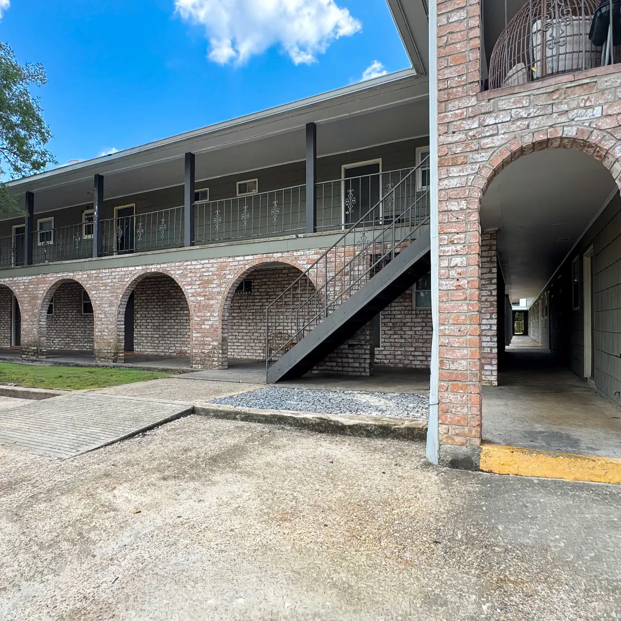 View of a brick apartment complex featuring arches and a staircase, with a grassy area nearby and blue sky with clouds.