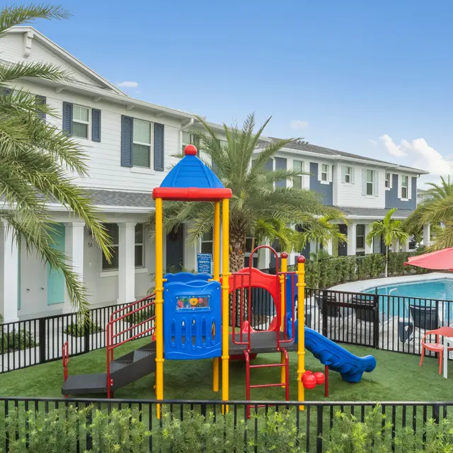 A colorful playground with slides and climbing structures surrounded by grass, located next to a swimming pool in a residential area with palm trees.