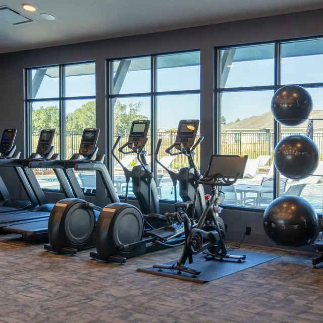 A modern fitness center featuring exercise equipment like treadmills, ellipticals, and a stationary bike, with large windows providing views of an outdoor pool area.