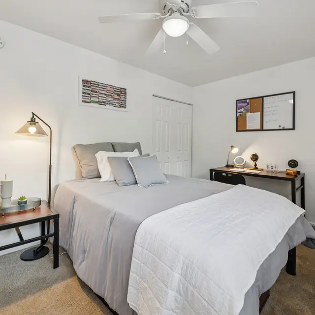 Cozy Bedroom Interior with Desk and Fan A cozy bedroom featuring a queen-sized bed with gray bedding, bedside table with lamp, a workspace desk, and a patterned pouf.