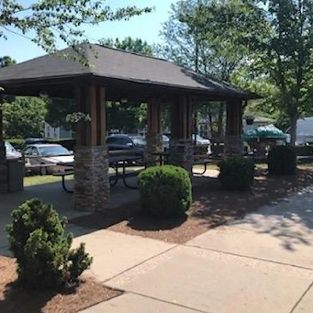 A park pavilion with a covered structure, surrounded by trees and bushes, with parked cars in the background on a sunny day.